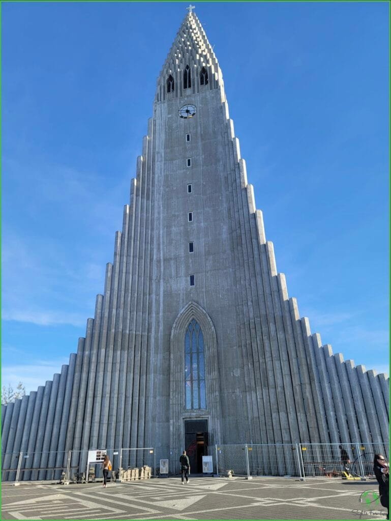 The image shows Hallgrímskirkja, a famous church in Reykjavik, Iceland. The church’s towering concrete facade is striking, with its unique stepped design that tapers towards the top, resembling volcanic basalt columns. A clock is visible near the top of the tower, and a small cross adorns the peak. The entrance is at the bottom center, with a few people walking towards it. The sky is clear and blue.