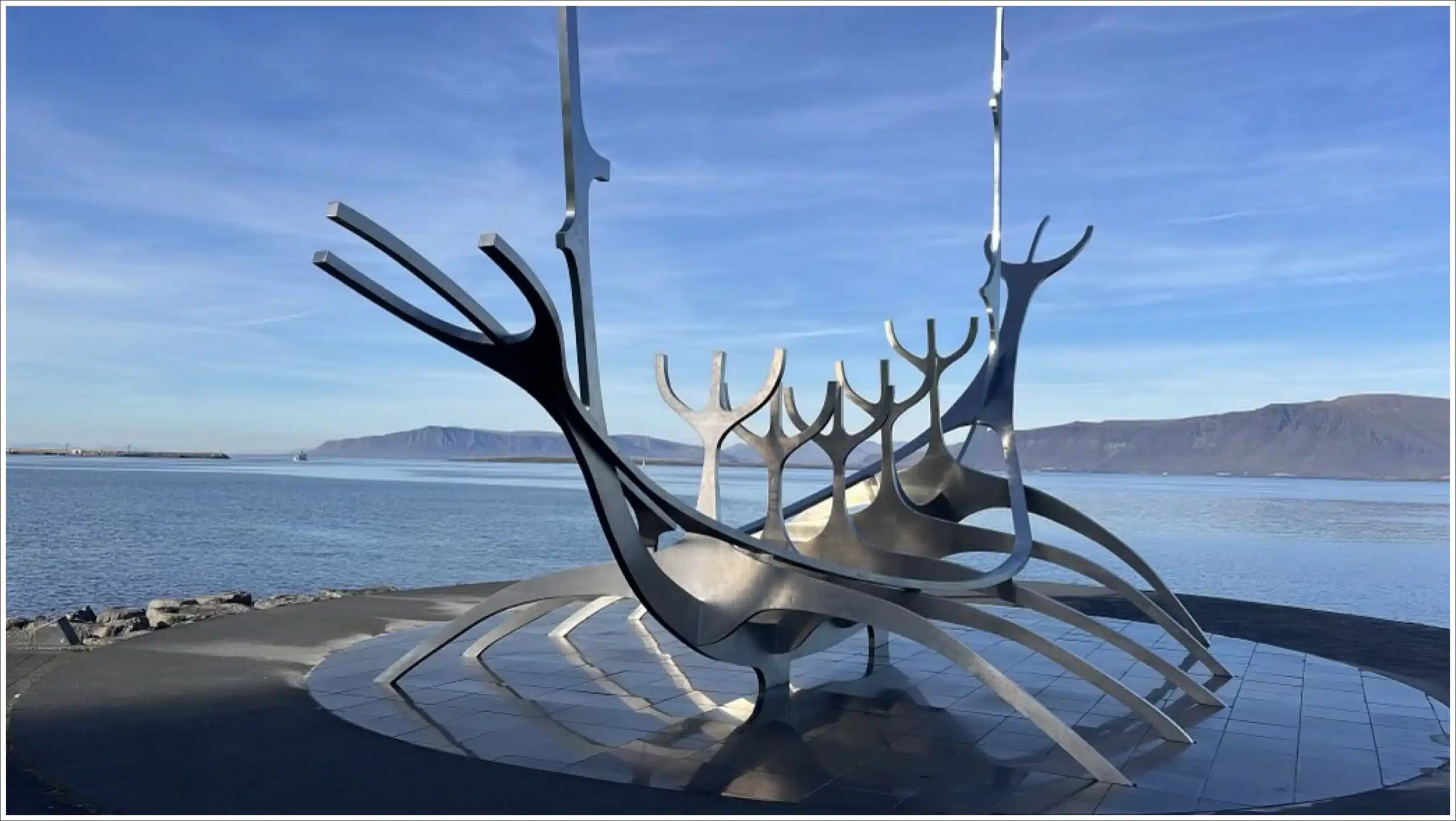 The image shows the Sun Voyager sculpture in Reykjavik, Iceland, a striking stainless steel structure resembling a Viking ship. The sculpture is set against a backdrop of calm water and distant mountains under a clear blue sky.